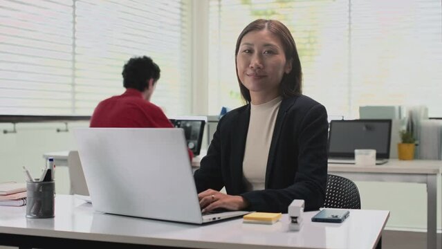 Medium portrait shot of female office worker texting messages to clients on laptop and looking at camera