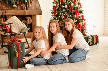 Obraz na płótnie Canvas Three pretty young girls in the living room are sitting on the floor near the Christmas tree. Christmas, New Year concept.