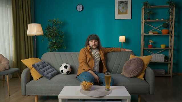 In the picture, a middle aged man is sitting leaning against the back of the sofa. He looks at the camera, next to him is a ball. On the table is a beer and chips. He looks unhappy, skeptical