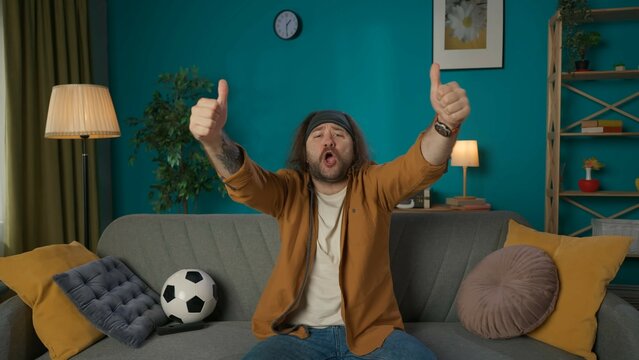 In the frame, a middle aged man is sitting on a couch in a room, with a soccer ball lying next to him. He is shown watching a soccer match, he is cheering, supporting, shouting. Depicts a fan