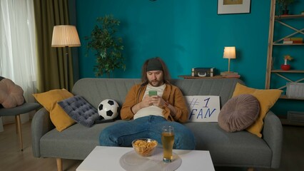 In the shot, a middle aged man is sitting on a couch, next to a soccer ball and a tablet. He is watching something on his phone. Playing a football fan, hes waiting for the match to start