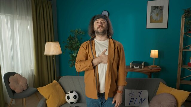 In the shot, a middle aged man stands in a room. With his hand over his heart he looks into the camera, miming the anthem. Next to him lies a soccer ball. He is impersonating a soccer fan