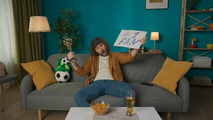 A middle aged man sits on a couch in a room. He is looking at the camera, yelling and holding a sign and a trophy. Imitates a soccer fan who supports the soccer team. Hes excited, waiting.