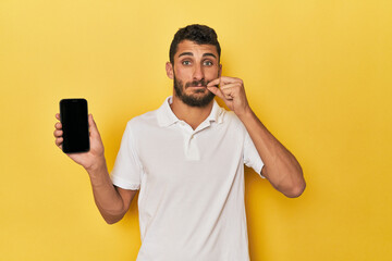 Young Hispanic man shows phone screen with fingers on lips keeping a secret.