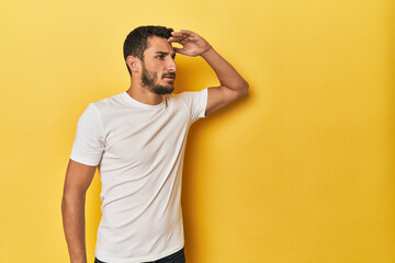 Young Hispanic man on yellow background looking far away keeping hand on forehead.