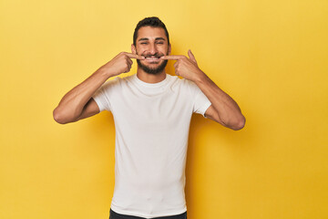 Young Hispanic man on yellow background smiles, pointing fingers at mouth.