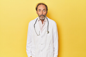 Doctor with stethoscope in yellow studio shrugs shoulders and open eyes confused.