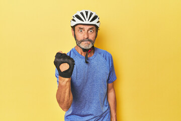 Cyclist wearing helmet in yellow studio showing fist to camera, aggressive facial expression.