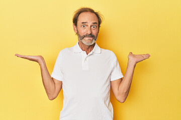 Caucasian middle-aged man in yellow studio confused and doubtful shrugging shoulders to hold a copy space.