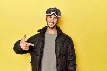Man in winter coat with snow goggles on yellow person pointing by hand to a shirt copy space, proud and confident