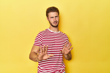 Young Caucasian man on a yellow studio background rejecting someone showing a gesture of disgust.