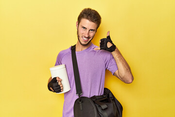 Man with protein jar and backpack on yellow showing a mobile phone call gesture with fingers.