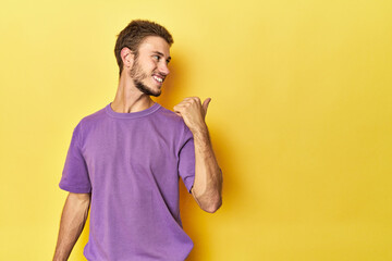 Young Caucasian man on a yellow studio background points with thumb finger away, laughing and carefree.