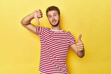 Young Caucasian man on a yellow studio background showing thumbs up and thumbs down, difficult...