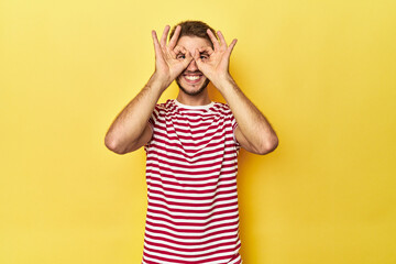 Young Caucasian man on a yellow studio background showing okay sign over eyes