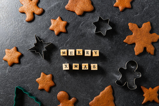 holidays, cooking and baking concept - close up of merry christmas words made of wooden toy blocks or stams, gingerbread cookies and molds on black table top at kitchen