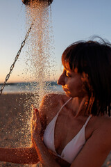 Beautiful young woman takes a shower on the beach near the sea. Attractive young woman in a white swimsuit enjoying a shower at sunset after swimming. close-up.