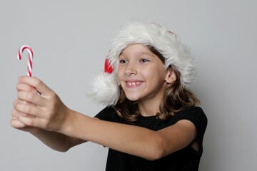 Portrait of a smiling girl wearing a Santa Claus hat with a striped Christmas candy. Funny playful child. New Year children's joy concept