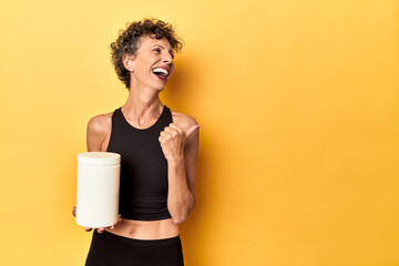 MIddle aged athlete woman holding protein supplement on yellow points with thumb finger away, laughing and carefree.
