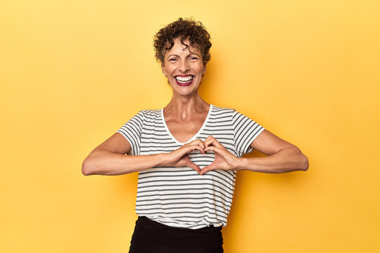 Mid-aged caucasian woman on vibrant yellow smiling and showing a heart shape with hands.