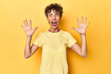 Mid-aged caucasian woman on vibrant yellow receiving a pleasant surprise, excited and raising hands.