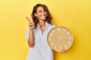 Middle aged woman holding a wall clock on a yellow backdrop joyful and carefree showing a peace...