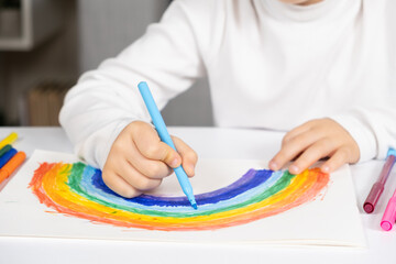 A small child draws a rainbow with felt-tip pens in a sketchbook while sitting at a table