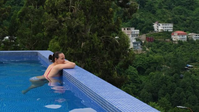 Young woman enjoys view while bathing in outdoor infinity pool, enjoying surrounding view, slow motion