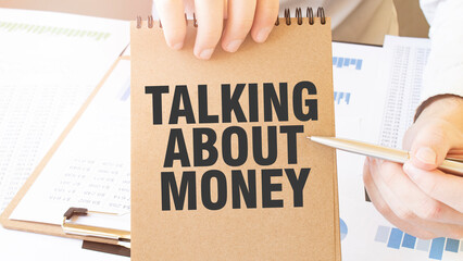 Text TALKING ABOUT MONEY on brown paper notepad in businessman hands on the table with diagram. Business concept - obrazy, fototapety, plakaty
