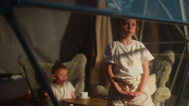 Upset girl looks out of window with younger brother in room. Bored little children tired of playing spend time at home. Unhappy kids in glamping cabin