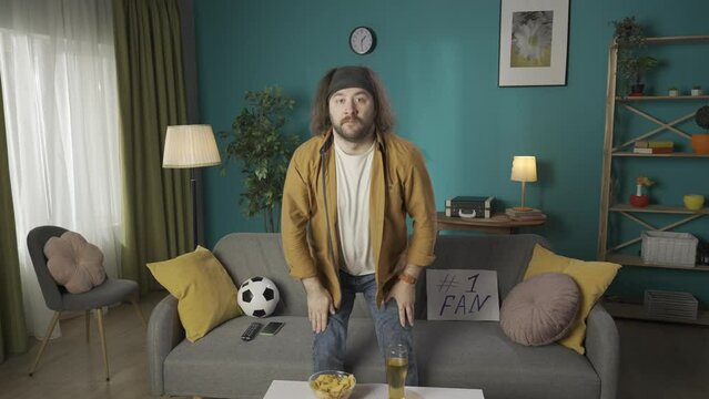 Footage shows a middle aged man sitting on a couch eating chips. Demonstrates watching a soccer match on television. He stands up puts his hand over his heart. Then sits down and eats chips HDR BT2020