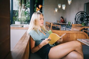 Fototapeta na wymiar Contemplative Caucasian student with education textbook for organization planning pondering on test information while doing homework in cafe interior, pensive blonde woman learning and studying