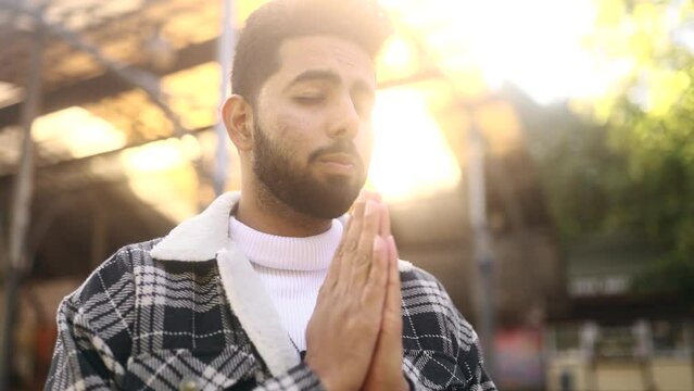 Portrait of hopeful young man clasping hands in prayer asking for blessing and help while the rays of rising sun fall on his face at city centre outdoors Religion and faith concept