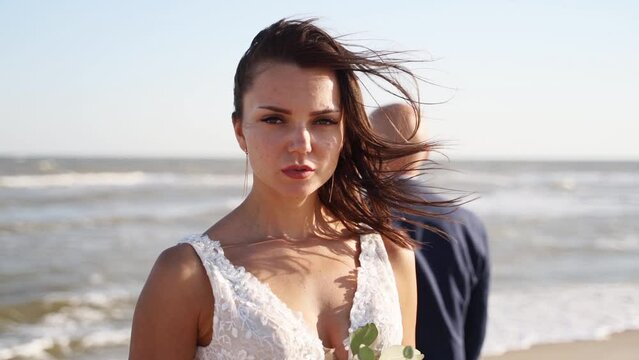 Pretty bride in gorgeous wedding dress looking at camera at the seaside, sea surf. Newlywed couple posing at the beach at wedding photoshoot. Woman, man standing at shore, windy weather.