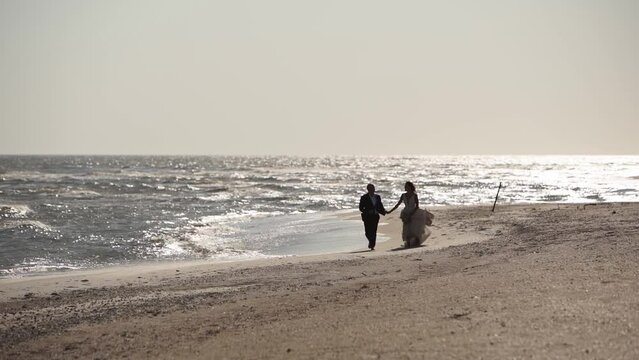 Just married couple holding hands, running at the sandy sea shore at wedding photoshoot. Happy bride with bouquet and groom having fun at the beach, gorgeous white dress waving on the wind.