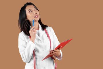 Thoughtful female Asian nutritionist with clipboard on beige background