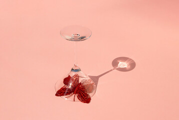 Red sparkly butterfly under a cocktail glass on sunny day. Creative concept on pink background.
