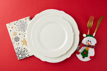 Make your Christmas meal unforgettable. Top view flat lay of snowman, crockery, napkin with pattern on red background