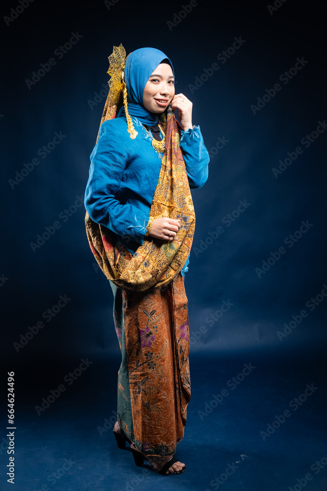 Wall mural Muslim woman wearing traditional wear blue kebaya and hijab isolated on blue background. Idul Fitri and hijab fashion concept.