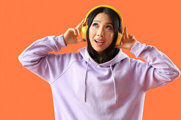 Young Asian woman in lilac hoodie with headphones on orange background