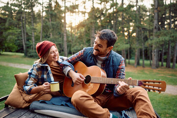 Happy man plays acoustic guitar to his girlfriend while camping in woods.
