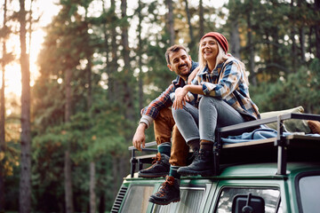 Happy couple sitting on top of their van while camping in woods.