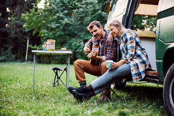 Cheerful man plays acoustic guitar to his girlfriend while camping in woods.