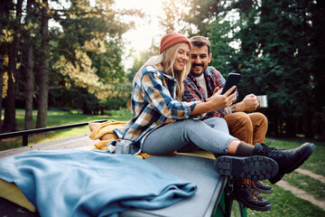 Happy couple taking selfie while sitting on roof of camping van in woods.