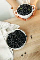 There are plates of ripe blueberries on the kitchen table and in the womans hands.