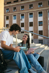 Young and happy college students with casual clothes sitting on stairs outdoors at university and talking while talking and studying with notes 