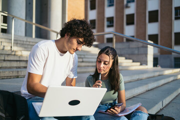 Young college students with casual clothes sitting on stairs outdoors at university and talking while working on laptop on class project