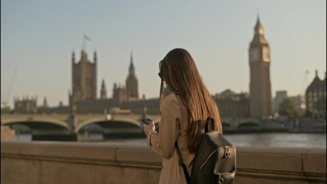 Young tourist woman using smartphone while walking along  Thames River with Big Ben Palace of Westminster background view, on a beautiful sunny morning
