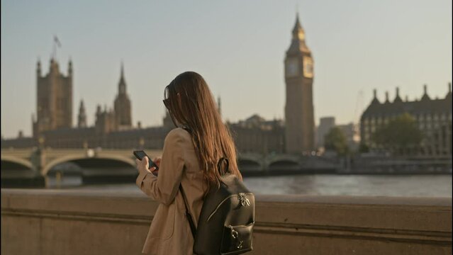 Young tourist woman using smartphone while walking along  Thames River with Big Ben Palace of Westminster background view, on a beautiful sunny morning
