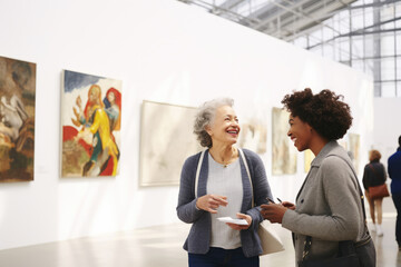 Females, adult woman and elegant lady visitors art gallery discussion artwork in exhibition at a modern museum of contemporary art. Abstract painting in the background on a white wall - obrazy, fototapety, plakaty